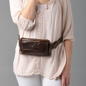 May include: A brown leather fanny pack with a rectangular shape and a front pocket. The belt is brown and has a buckle closure. The fanny pack is worn around the waist.