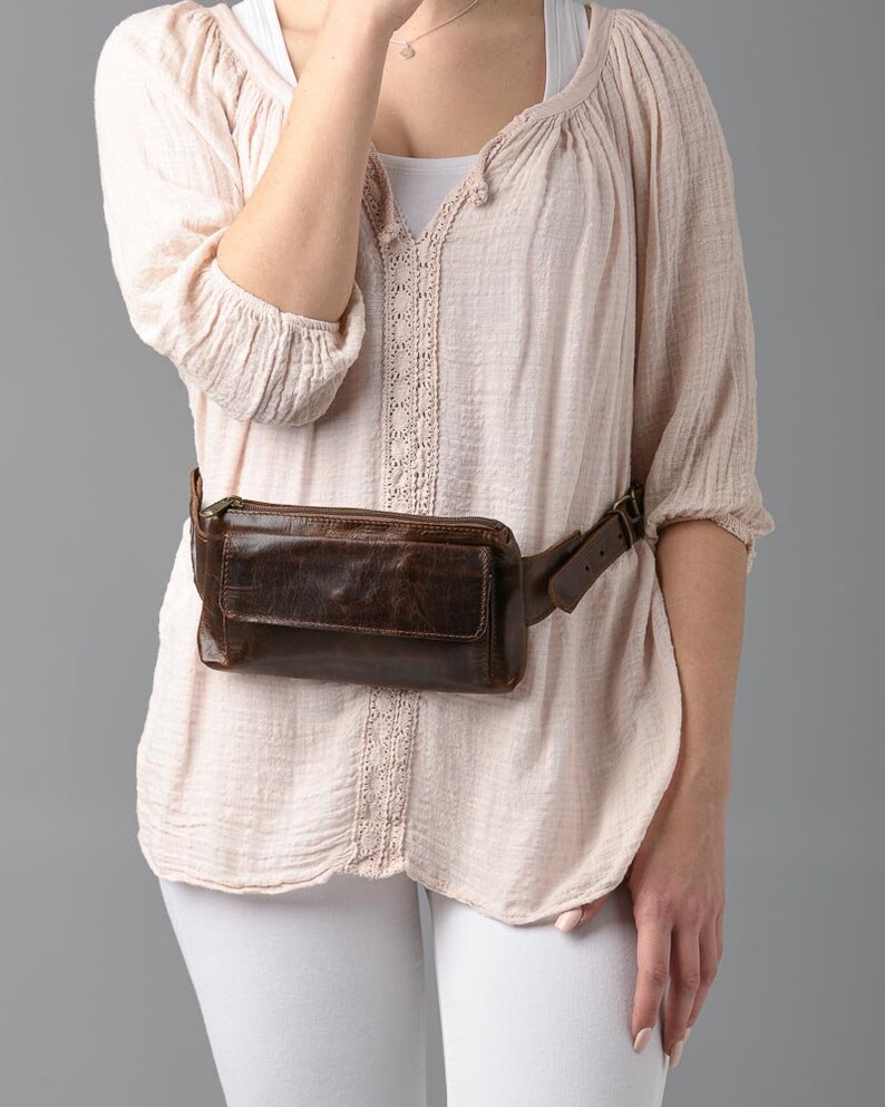 May include: A brown leather fanny pack with a rectangular shape and a front pocket. The belt is brown and has a buckle closure. The fanny pack is worn around the waist.