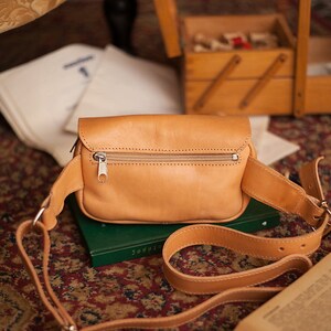 Peut inclure: Une banane en cuir de couleur tan avec une fermeture &agrave; glissi&egrave;re et une sangle r&eacute;glable. Le sac est pos&eacute; sur un livre vert.