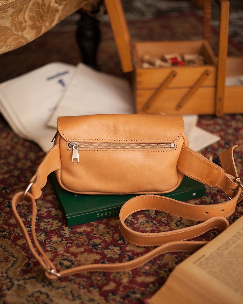 Peut inclure: Une banane en cuir de couleur tan avec une fermeture &agrave; glissi&egrave;re et une sangle r&eacute;glable. Le sac est pos&eacute; sur un livre vert.