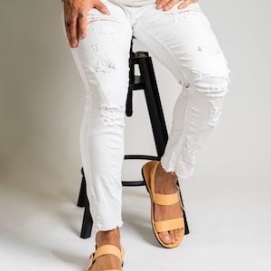 May include: White denim jeans featuring a distressed, ripped design with frayed hems at the ankles, paired with light tan leather sandals that have two horizontal straps and a side buckle closure, worn with a plain light-colored t-shirt.
