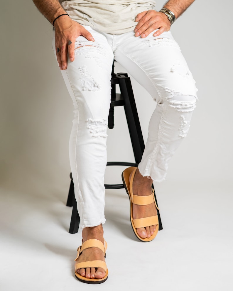May include: White denim jeans featuring a distressed, ripped design with frayed hems at the ankles, paired with light tan leather sandals that have two horizontal straps and a side buckle closure, worn with a plain light-colored t-shirt.