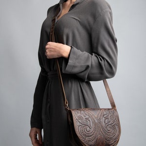 Peut inclure: Un sac &agrave; bandouli&egrave;re en cuir marron avec des sculptures florales complexes. Le sac a une fermeture &agrave; rabat et un d&eacute;tail de boucle.