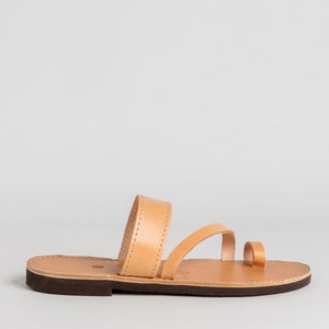 May include: A tan leather sandal featuring a wide strap across the midfoot, a thinner strap extending toward the toes, and a separate toe loop. The sandal has a flat, dark brown sole with visible stitching along the edges of the leather straps.