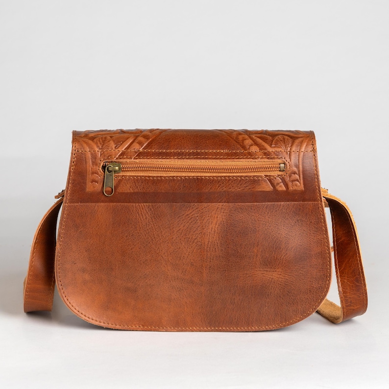 May include: A brown leather crossbody bag with a flap closure and a long adjustable strap. The bag features a zipper pocket and decorative embossed detailing on the flap. The bag is made of a smooth, durable leather.