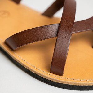 May include: A brown leather sandal featuring thin, crisscrossing straps attached to a light tan leather footbed. The sandal has a visible white stitched border along the edge of the sole and a solid black rubber base layer for the bottom of the shoe.