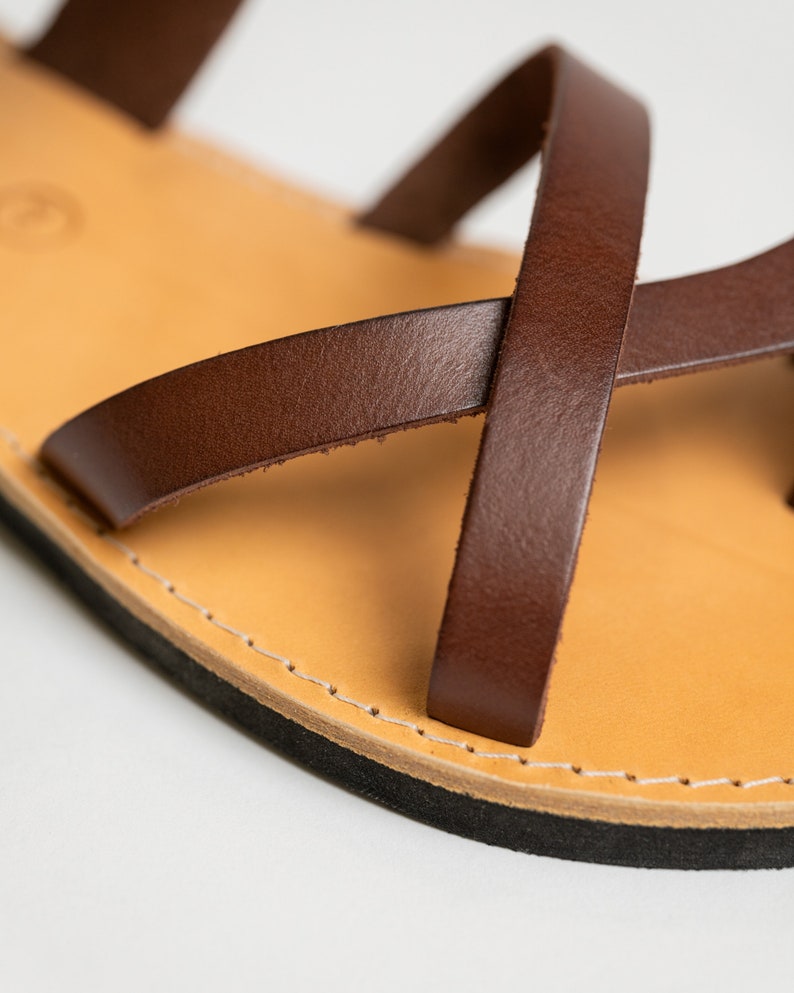 May include: A brown leather sandal featuring thin, crisscrossing straps attached to a light tan leather footbed. The sandal has a visible white stitched border along the edge of the sole and a solid black rubber base layer for the bottom of the shoe.