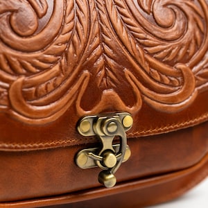 May include: Close-up of a brown leather bag with intricate floral and leaf carvings. The bag features a brass clasp and a detailed embossed design. The leather has a rich, warm tone.
