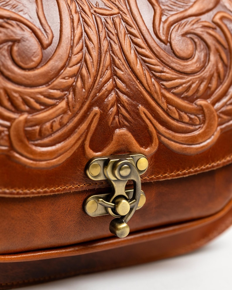 May include: Close-up of a brown leather bag with intricate floral and leaf carvings. The bag features a brass clasp and a detailed embossed design. The leather has a rich, warm tone.