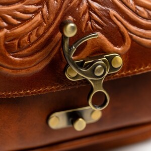 May include: Close-up of a brown leather bag with intricate embossed floral patterns. The bag features a brass clasp and hardware. The leather has a rich, warm tone, and the design suggests a vintage or handcrafted style.