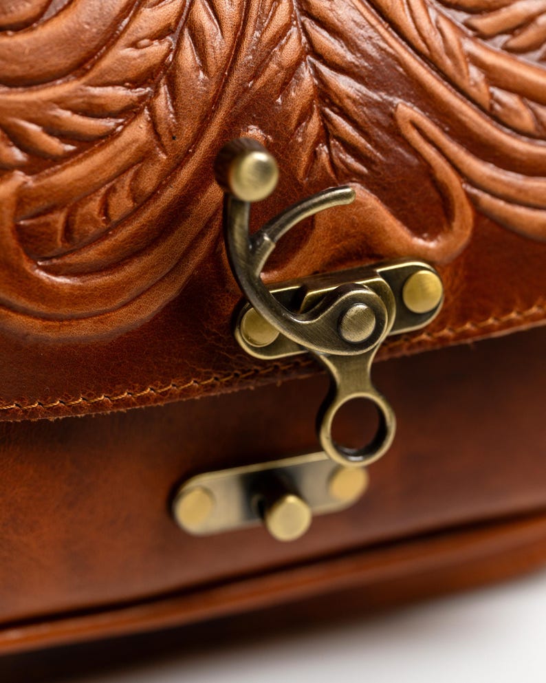May include: Close-up of a brown leather bag with intricate embossed floral patterns. The bag features a brass clasp and hardware. The leather has a rich, warm tone, and the design suggests a vintage or handcrafted style.