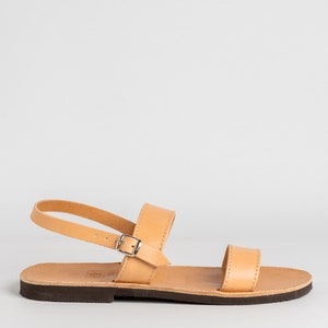 May include: A tan leather sandal featuring a flat dark brown sole, a single horizontal strap across the toes, and an adjustable ankle strap secured with a small silver-toned metal buckle. The design includes visible stitching along the edges.