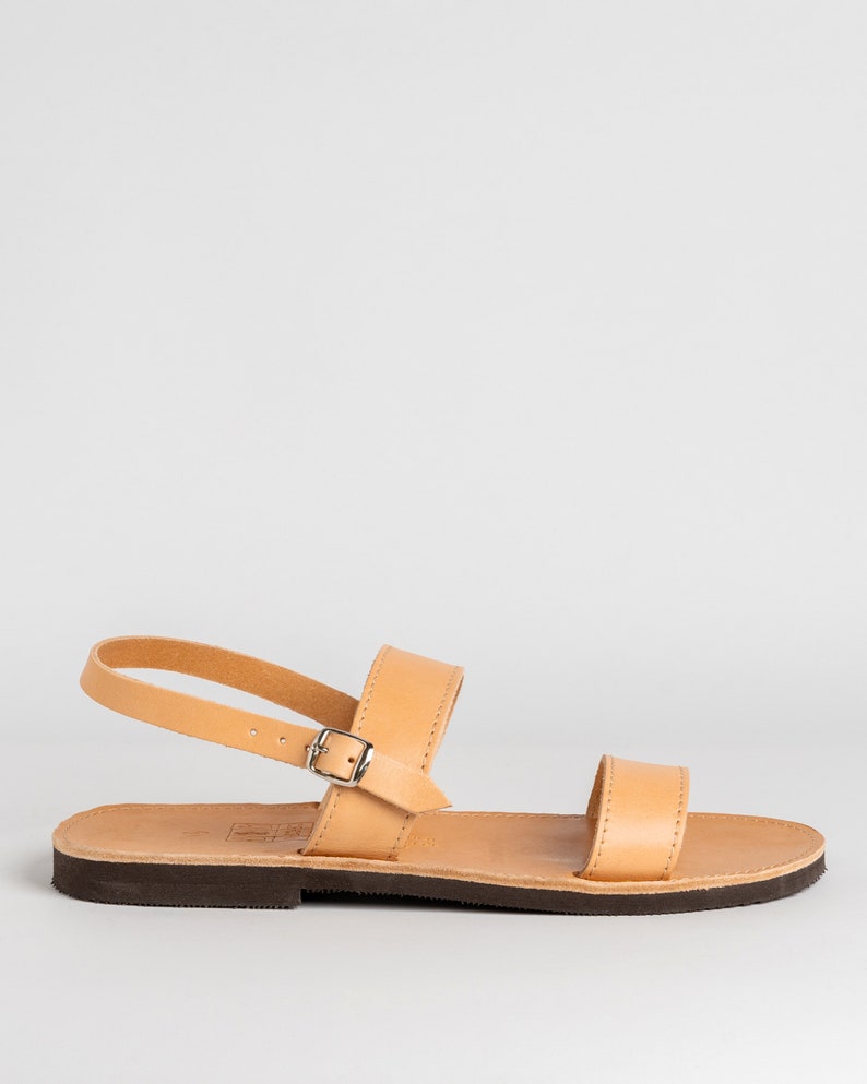 May include: A tan leather sandal featuring a flat dark brown sole, a single horizontal strap across the toes, and an adjustable ankle strap secured with a small silver-toned metal buckle. The design includes visible stitching along the edges.