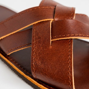 May include: A brown leather sandal featuring a crisscross strap design with visible stitching along the edges. The sandal has a smooth, dark brown footbed and a textured black sole, showcasing the natural grain and texture of the leather material.