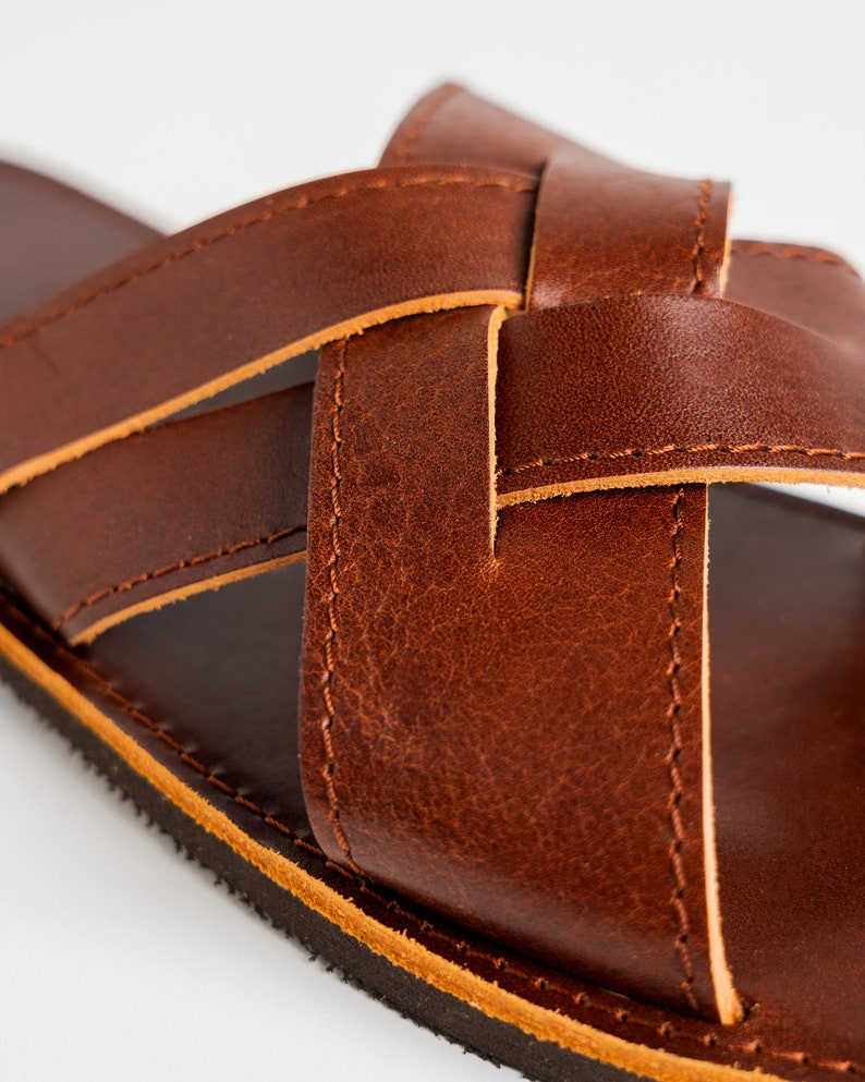 May include: A brown leather sandal featuring a crisscross strap design with visible stitching along the edges. The sandal has a smooth, dark brown footbed and a textured black sole, showcasing the natural grain and texture of the leather material.
