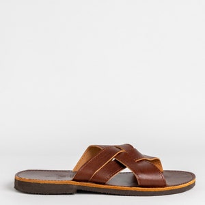 May include: A brown leather slide sandal featuring a wide, crisscross strap design across the top. The sandal has a flat, dark brown sole with a visible tan-colored midsole layer and a smooth, polished finish on the upper leather straps.