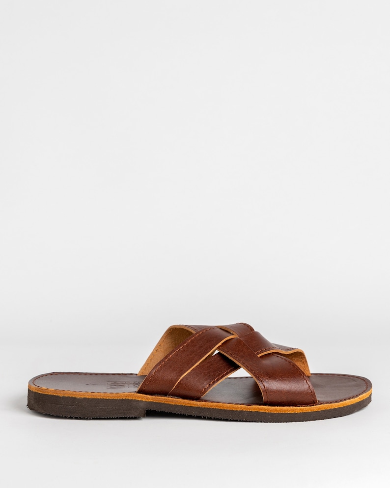 May include: A brown leather slide sandal featuring a wide, crisscross strap design across the top. The sandal has a flat, dark brown sole with a visible tan-colored midsole layer and a smooth, polished finish on the upper leather straps.