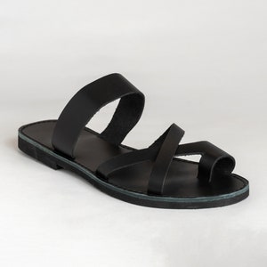May include: A black flat sandal featuring a wide strap across the midfoot and a thinner strap that crosses over the top of the foot, looping around the big toe. The sandal has a simple, low-profile sole and a smooth, matte finish throughout.