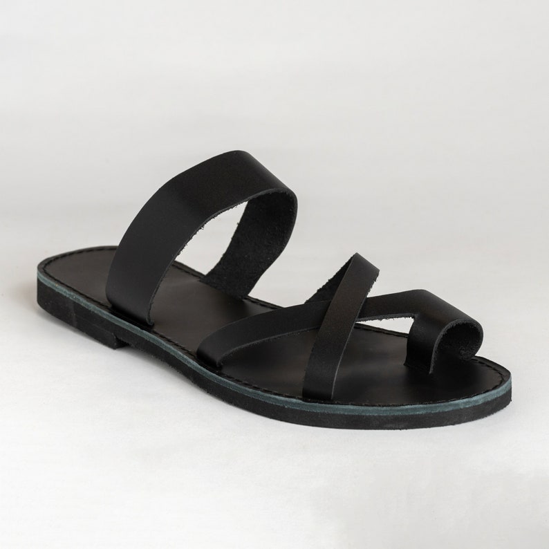 May include: A black flat sandal featuring a wide strap across the midfoot and a thinner strap that crosses over the top of the foot, looping around the big toe. The sandal has a simple, low-profile sole and a smooth, matte finish throughout.
