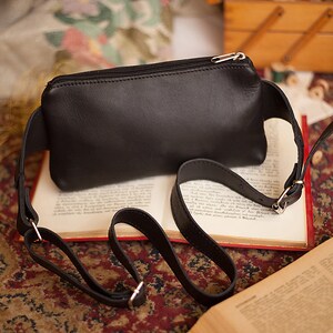 May include: A black leather fanny pack with a silver zipper closure and adjustable strap. The bag is rectangular and sits on an open book with red edges. The strap is attached with silver metal hardware.