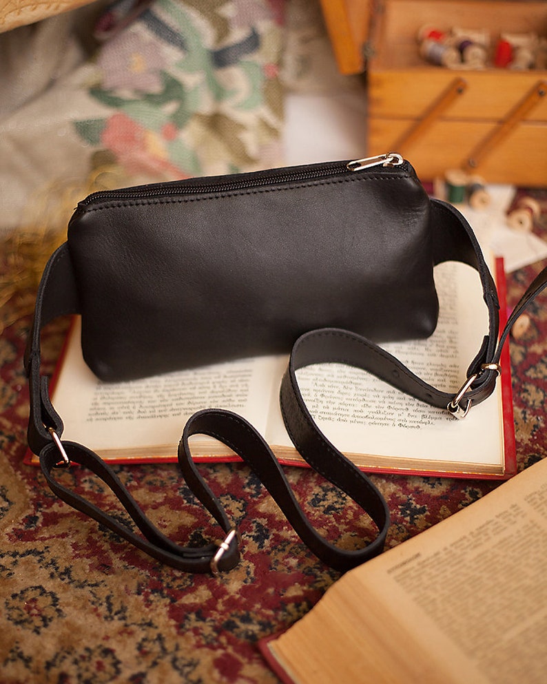 May include: A black leather fanny pack with a silver zipper closure and adjustable strap. The bag is rectangular and sits on an open book with red edges. The strap is attached with silver metal hardware.