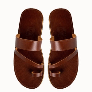 May include: A pair of dark brown leather sandals featuring a wide horizontal strap across the midfoot and a separate loop for the big toe. The footbed is dark brown with a visible embossed icon stamp near the heel and a thin light-colored sole edge.