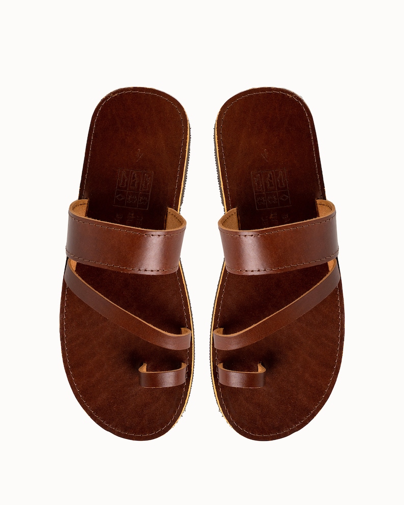 May include: A pair of dark brown leather sandals featuring a wide horizontal strap across the midfoot and a separate loop for the big toe. The footbed is dark brown with a visible embossed icon stamp near the heel and a thin light-colored sole edge.