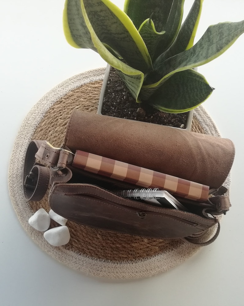 Puede incluir: Un bolso de cuero marr&oacute;n con cierre de cremallera. El bolso est&aacute; abierto y contiene un cuaderno, una libreta de espiral y una libreta de notas de madera.