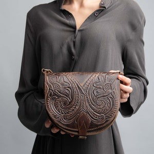May include: A brown leather crossbody bag with intricate floral carvings. The bag has a flap closure with a buckle and a strap that can be adjusted.
