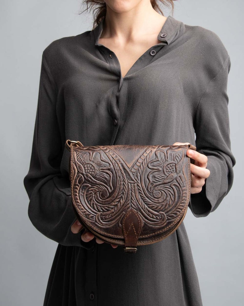 May include: A brown leather crossbody bag with intricate floral carvings. The bag has a flap closure with a buckle and a strap that can be adjusted.