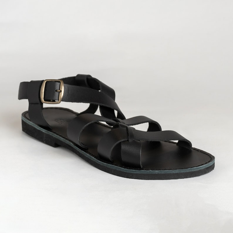 May include: A black leather sandal featuring multiple wide straps across the foot and an adjustable ankle strap with a metallic brass-toned buckle. The footwear has a flat, dark sole and a simple, open-toe design with a secure heel enclosure.