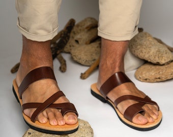 May include: Brown leather sandals with criss-cross straps and a thong toe. The sandals are being worn by a person standing on a rock.