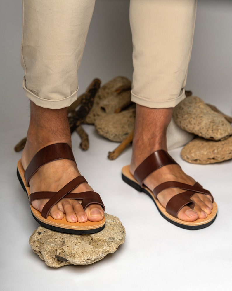 May include: A pair of dark brown leather sandals featuring a wide strap across the instep and thinner straps that cross over the top of the foot, including a toe loop. The sandals have a tan leather footbed and a thin black rubber outsole.