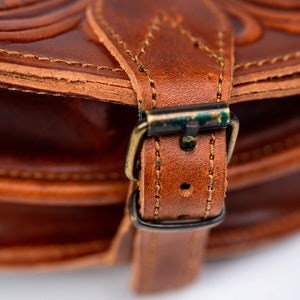 May include: Close-up of a brown leather bag with a brass buckle. The bag has a decorative embossed pattern and stitching.