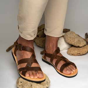 May include: A pair of brown leather sandals featuring multiple crisscrossing straps across the top of the foot, a secure ankle strap with a metal buckle, a light tan leather footbed, and a thin black rubber sole for traction and durability.
