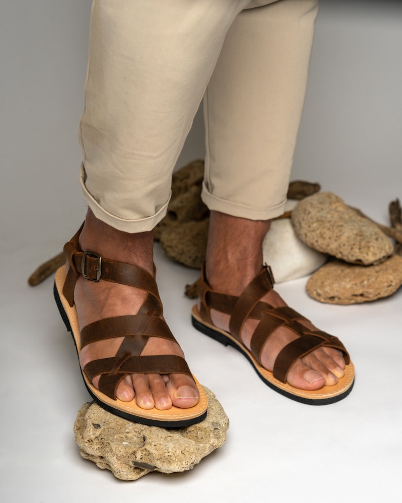 May include: A pair of brown leather sandals featuring multiple crisscrossing straps across the top of the foot, a secure ankle strap with a metal buckle, a light tan leather footbed, and a thin black rubber sole for traction and durability.