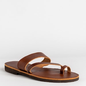 May include: A brown leather slide sandal featuring a toe loop and a wide strap across the midfoot. The sandal has a flat, dark brown sole with visible stitching along the edge of the footbed and a smooth, matte finish on the leather straps.