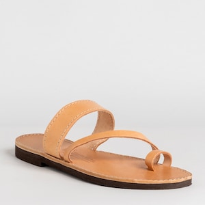 May include: A tan leather sandal featuring a flat dark brown sole, a wide strap across the midfoot, and a separate loop for the big toe. The natural leather upper is secured with light-colored stitching along the edges of the straps and footbed.