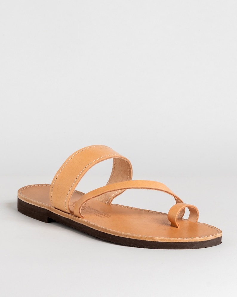May include: A tan leather sandal featuring a flat dark brown sole, a wide strap across the midfoot, and a separate loop for the big toe. The natural leather upper is secured with light-colored stitching along the edges of the straps and footbed.