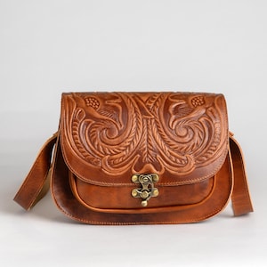 May include: A brown leather crossbody bag with a detailed floral embossed design on the flap. The bag features a brass clasp closure and an adjustable shoulder strap. The bag is approximately 10 inches wide.