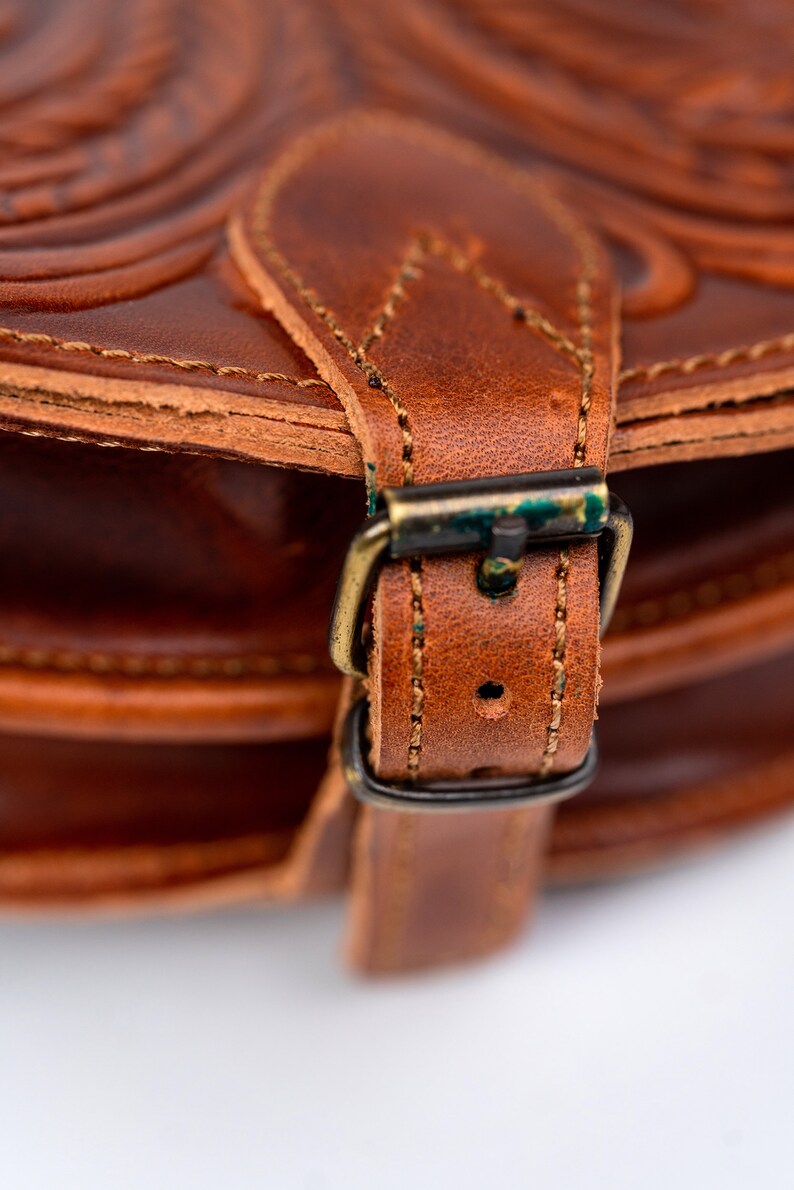 Puede incluir: Primer plano de un bolso de cuero marr&oacute;n con una hebilla de lat&oacute;n. El bolso tiene un patr&oacute;n decorativo en relieve y costuras.