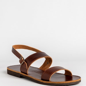 May include: A dark brown leather sandal featuring two wide straps across the top, an adjustable ankle strap with a small metal buckle, and a flat dark brown sole. The inner sole is stamped with text that reads Real Leather Made in Brazil.