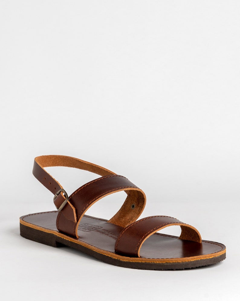 May include: A dark brown leather sandal featuring two wide straps across the top, an adjustable ankle strap with a small metal buckle, and a flat dark brown sole. The inner sole is stamped with text that reads Real Leather Made in Brazil.
