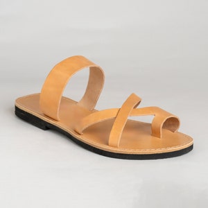 May include: A tan leather sandal featuring a wide arch strap and a crisscross toe loop design. The sandal has a flat, black rubber sole with visible white stitching along the edge, providing a simple and functional construction for this footwear.