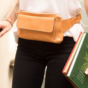 May include: A tan leather fanny pack with a front pocket and a zipper closure. The adjustable belt is fastened around the waist. The person is wearing black jeans and a white top. A book with a green cover is held in the hand.