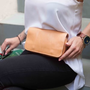 Peut inclure: Une banane en cuir marron clair avec une fermeture &eacute;clair. La banane est port&eacute;e autour de la taille d'une personne portant un d&eacute;bardeur blanc et un jean noir.