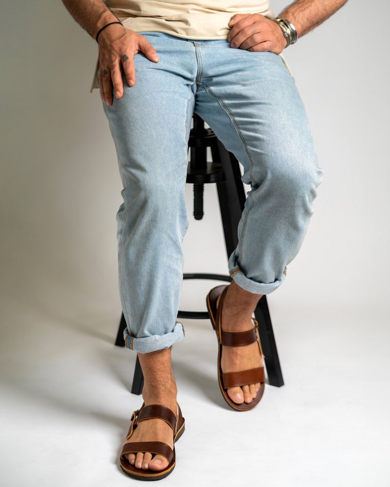 May include: Light blue denim jeans with a straight-leg cut and cuffed hems, paired with brown leather sandals featuring two horizontal straps and a side buckle closure. The jeans have a classic five-pocket design and a light wash finish.