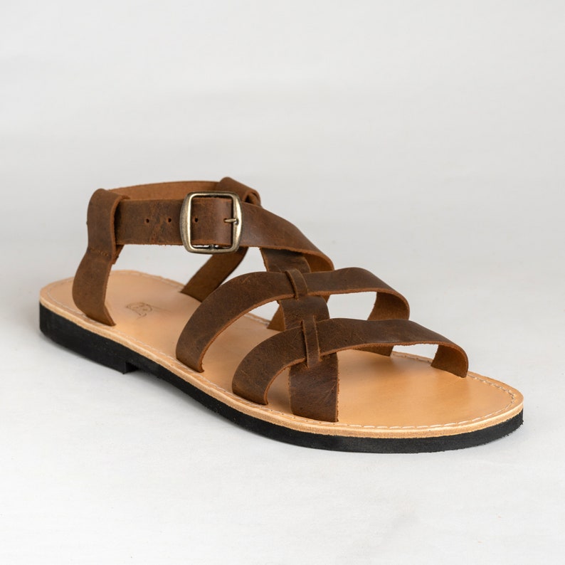 May include: A brown leather sandal featuring multiple crisscrossing straps across the top and an adjustable ankle strap with a brass-toned metal buckle. The sandal has a light tan leather footbed and a flat, black rubber sole with visible stitching.
