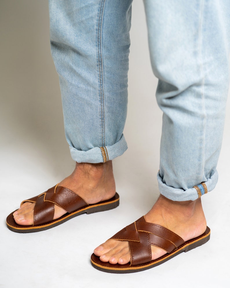 May include: A pair of brown leather slide sandals featuring a crisscross strap design across the top of the foot. The sandals have a flat, dark brown sole and an open-toe construction, providing a simple and functional footwear option.