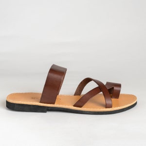 May include: A dark brown leather sandal featuring a wide strap across the midfoot and a crisscross strap design at the front with a toe loop. The sandal has a tan leather insole with visible stitching along the edge and a flat black rubber sole.